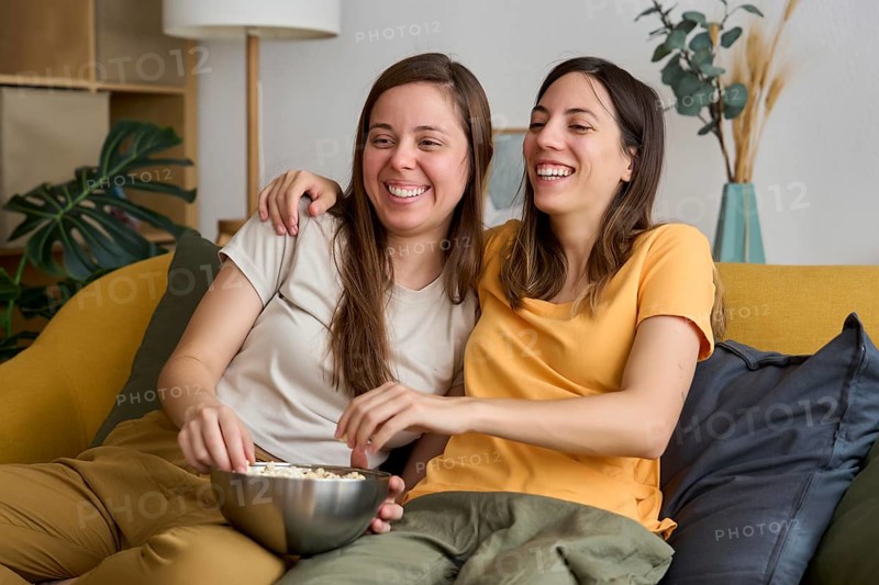 A happy lesbian couple sits comfortably on the couch, sharing a bowl of popcorn and laughing together The image captures a warm moment of love and companionship
