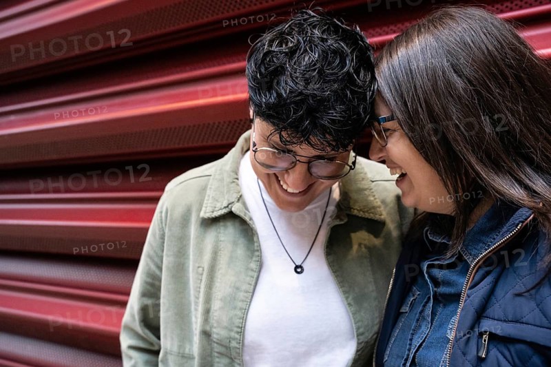 A joyful lesbian couple shares a laugh against a bright red backdrop. Their affectionate embrace captures the essence of love and happiness in an urban setting