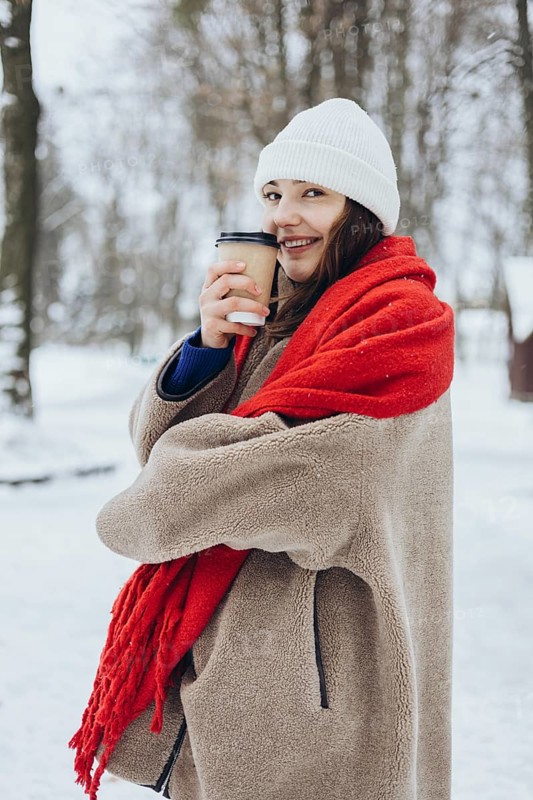 A cheerful young woman wrapped in a cozy coat and a red scarf, holding a coffee cup in a snowy park