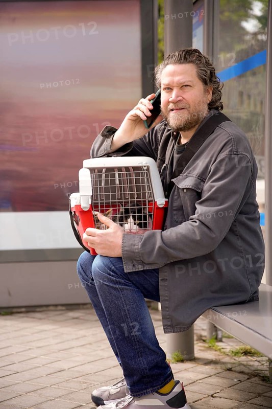 A man sits at a bus stop holding a cat carrier, engaged in conversation on his phone. The scene depicts a moment of routine as he waits, with his pet safely by his side