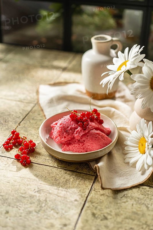 From above freshly made pink berry mousse garnished with red currants, placed in a ceramic bowl beside a vase with white daisies on a tiled table