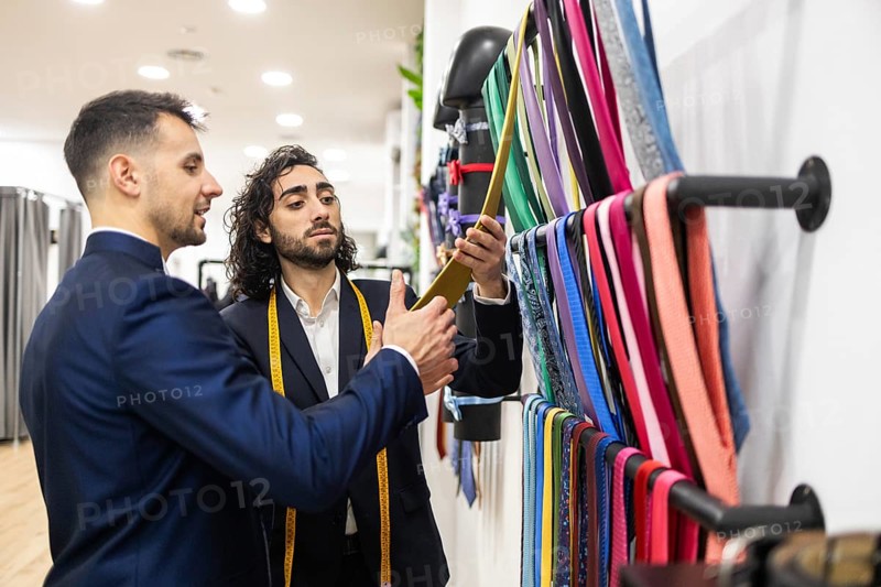 A tailor helps a client select a tie in a suit men store. The professional advises the customer on color and style, surrounded by a variety of elegant neckties