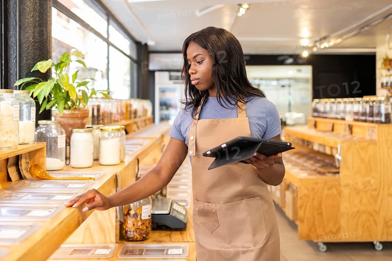 A dedicated store clerk checks inventory and organizes products in a vibrant organic bulk store. With a background of shelves, she holds a tablet to manage the inventory efficiently