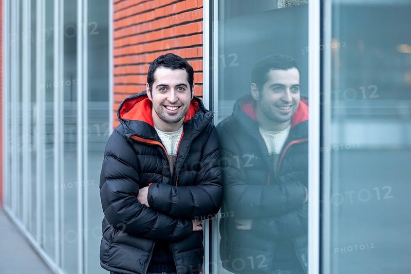 A man in a warm jacket leans against a glass wall, his reflection visible beside him. The setting at a mental health leisure center highlights confidence and positivity in a supportive environment
