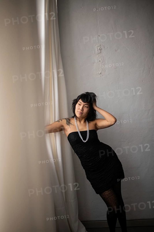 A graceful Asian woman poses by a textured wall, wearing a strapless black dress and pearl necklace. Her confident expression and stylish attire exude elegance