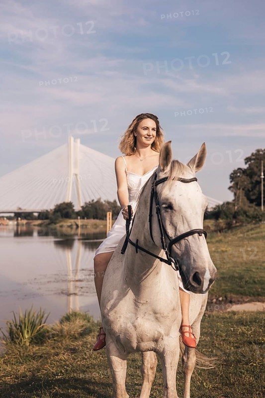 A young woman in a white dress rides a white horse beside a shimmering river. A striking cable stayed bridge frames the scene under a clear, sunny sky