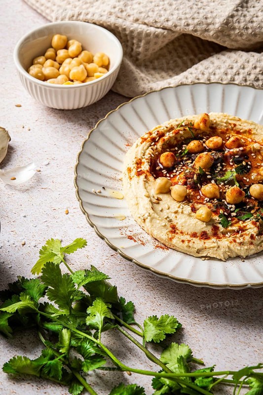 A plate of creamy hummus garnished with chickpeas, herbs, and spices sits on a textured background. A bowl of chickpeas and fresh herbs enhance the vibrant presentation