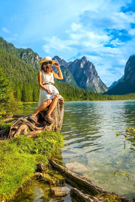 Young woman with straw hat sitting on a tree trunk by the lake admiring the mountain landscape in the dolomites