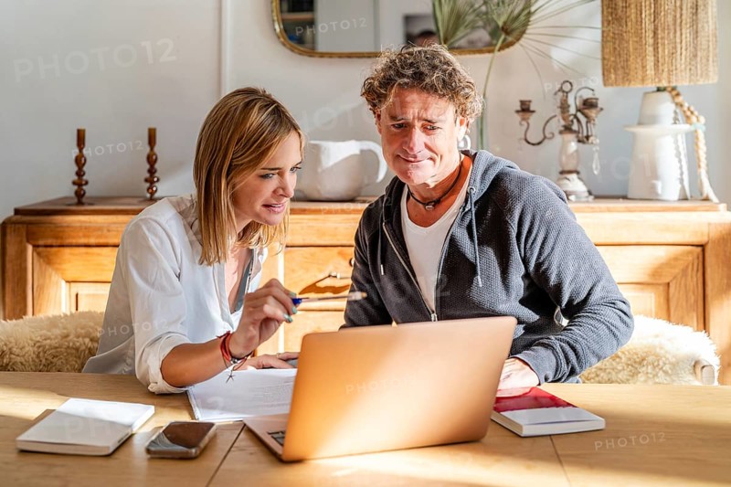 A couple engaged in work at home, sitting at a wooden table with a laptop and documents The room has warm lighting, creating a cozy and productive atmosphere