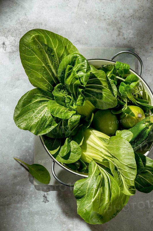 A colander filled with fresh green vegetables, including bok choy, lime, and zucchini, is placed on a rustic kitchen table. The natural sunlight enhances their vibrant colors
