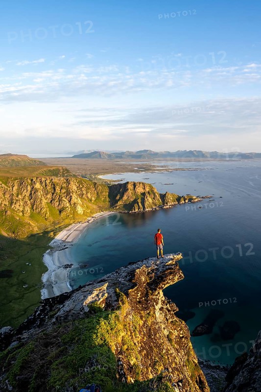 Evening mood, mountaineer standing on a rocky outcrop on the summit of Måtind, view of coastal landscape, Høyvika beach and sea, Måtinden, Andøya island, Vesterålen, Nordland Norway