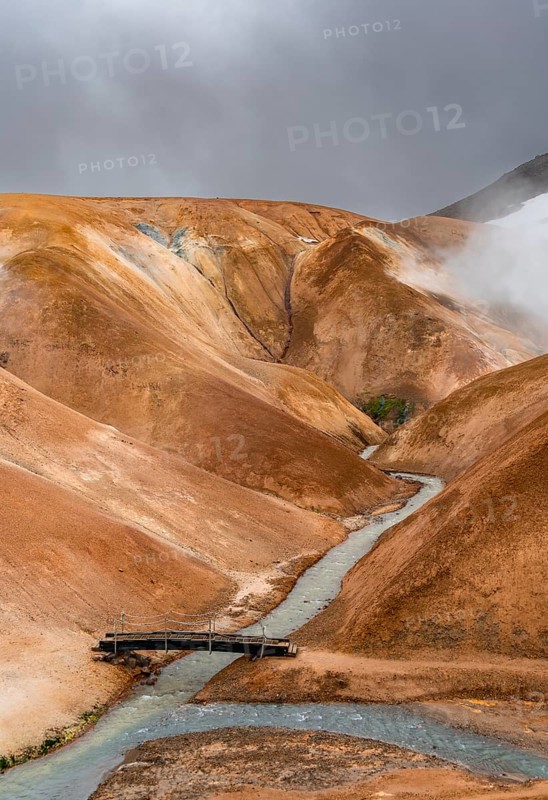 Steaming stream and bridge between colourful rhyolite mountains with snowfields, Hveradalir geothermal area, Kerlingarfjöll, Icelandic Highlands, Iceland