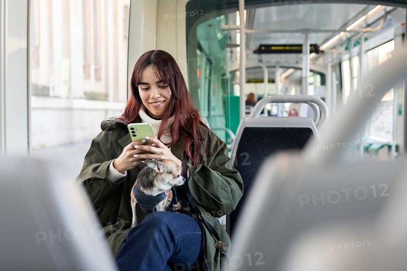 A young woman with a smartphone and a small chihuahua sits in a modern tram, enjoying a comfortable commute. She wears a casual outfit, reflecting urban lifestyle trends