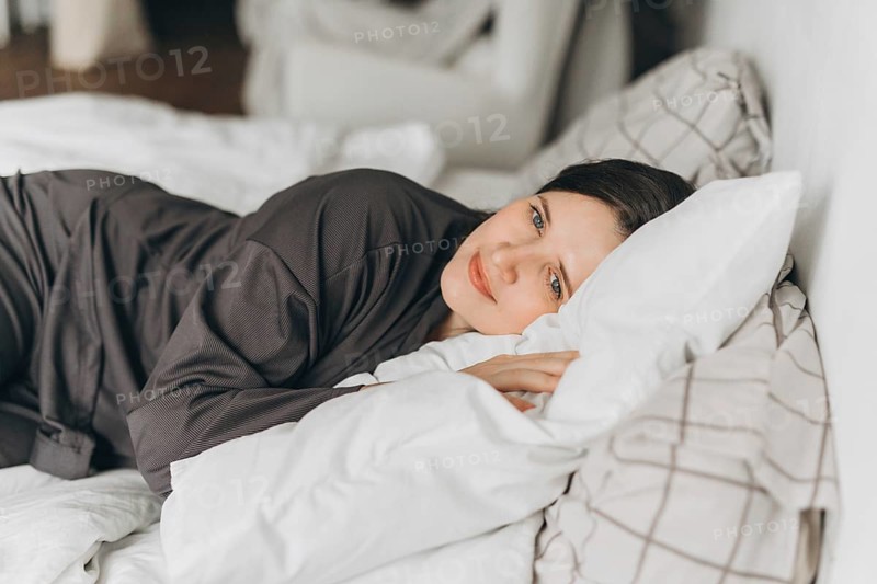A young woman in comfortable loungewear lies on a white bed, resting her head on soft pillows while smiling gently. The warm and cozy atmosphere highlights relaxation and comfort
