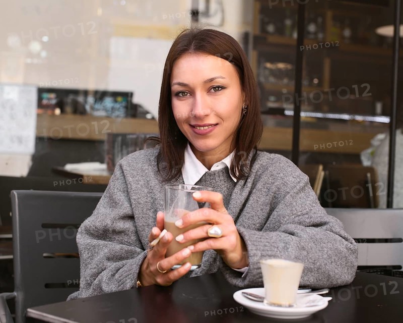 A woman in a gray sweater sits at a bar table, holding a cup of coffee. She smiles warmly, exuding a relaxed and content vibe in an inviting bar setting