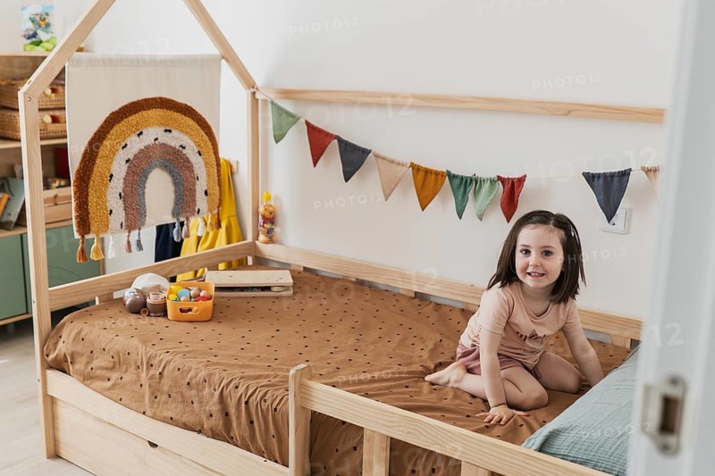 A happy young girl sits in a cozy bedroom with colorful decor, including a rainbow tapestry and vibrant bunting. The space is warm and playful, ideal for a big sister