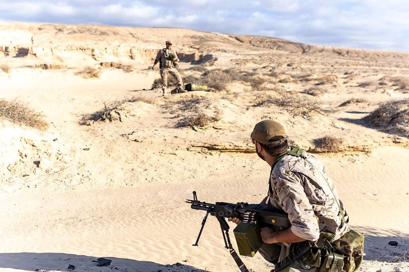Two soldiers in camouflage gear conduct training exercises in a desert environment. One soldier is holding a rifle, demonstrating military readiness and tactics