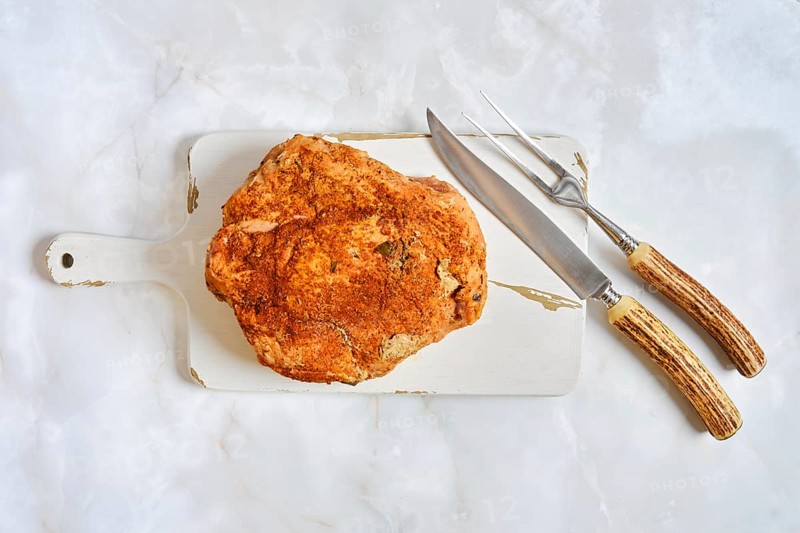 An oven roasted pork shoulder on a cutting board. Two carving tools sit beside it. The setting has a light marble surface