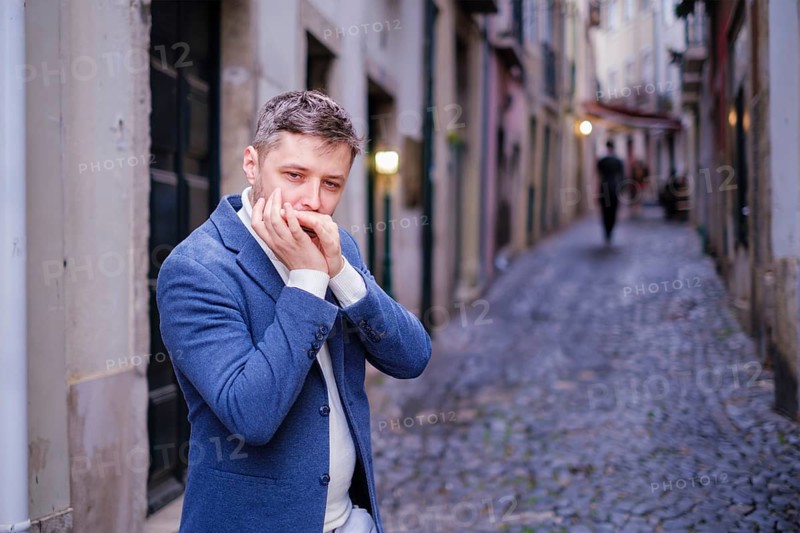 Man musician in a blue blazer and white sweater playing blues on a harmonica with eyes closed, standing outdoors in city street