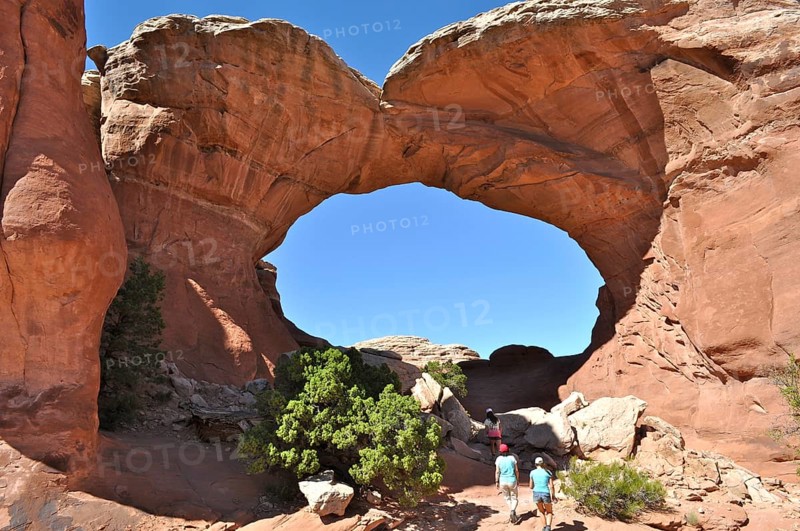 Group of hikers under a large rock arch, the Broken Arch, on a sunny day, Arches National Park, Utah, USA