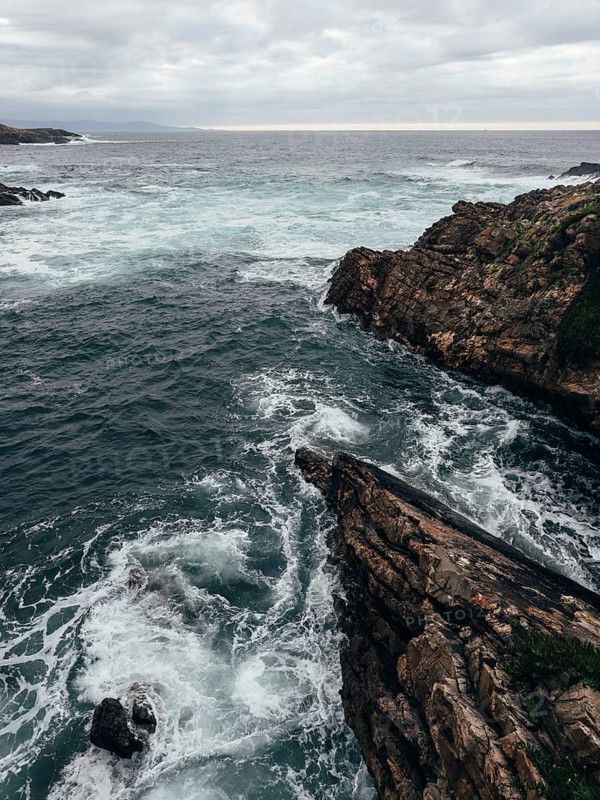 The image captures a dramatic coastal landscape with rocky cliffs and turbulent waves crashing against them. The overcast sky adds to the moody atmosphere, emphasizing the power and beauty of nature