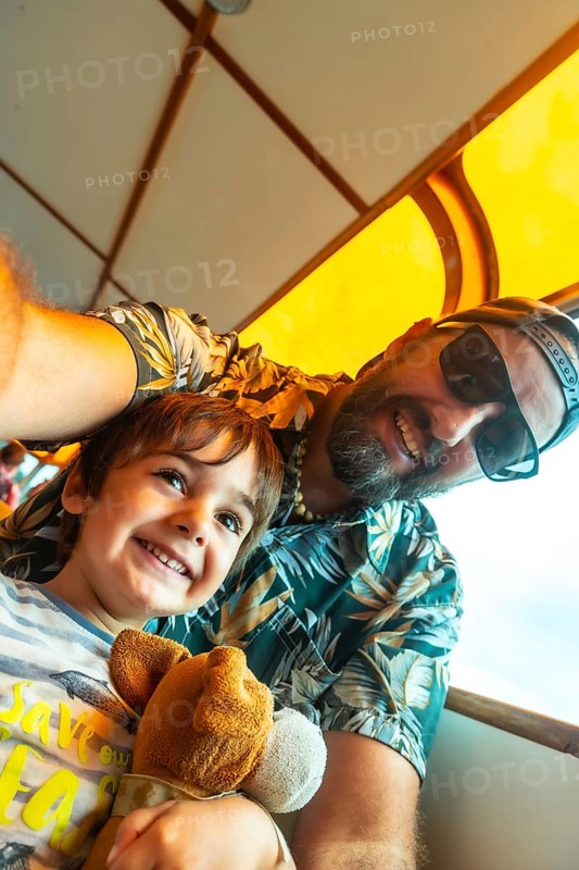 A man and a child are smiling at the camera. The man is wearing a green shirt with palm trees on it. The child is holding a stuffed animal