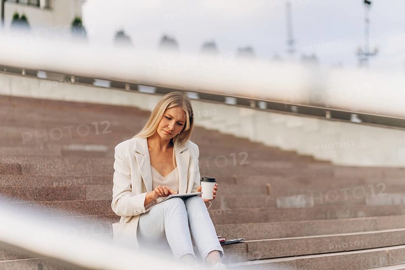 A businesswoman in a light blazer sits on outdoor steps, holding a tablet and a coffee cup. The setting hints at business, blending relaxation and focus