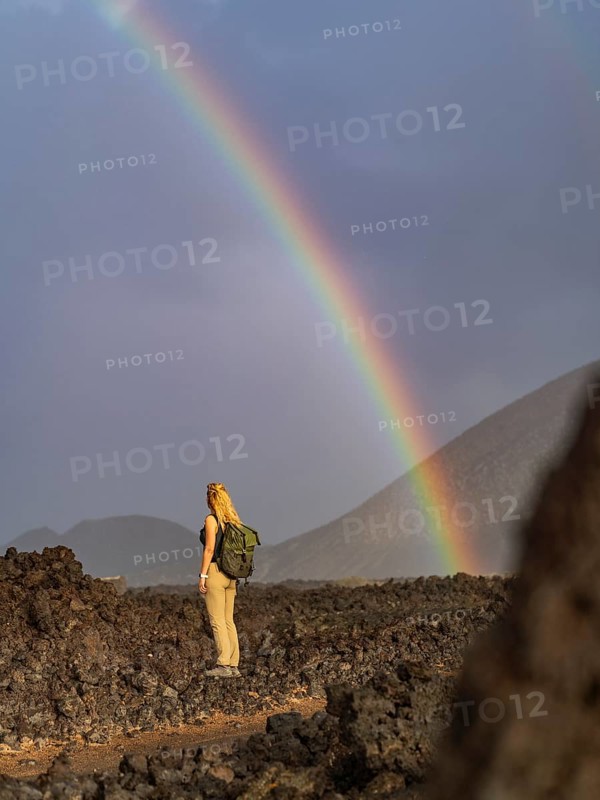 A woman with a backpack stands on volcanic terrain in Timanfaya National Park, Lanzarote, Spain, gazing at a vivid rainbow after rainfall, creating a striking natural scene