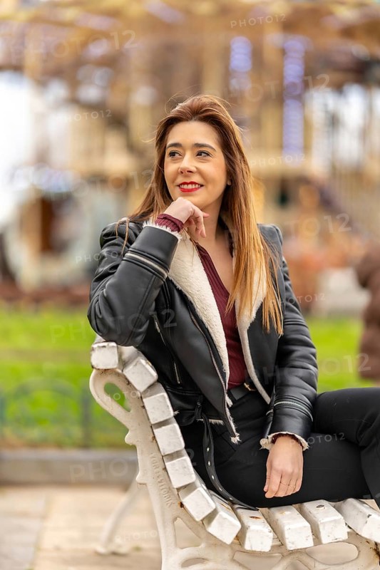 Young woman wearing a black jacket is smiling while sitting on a bench in an amusement park