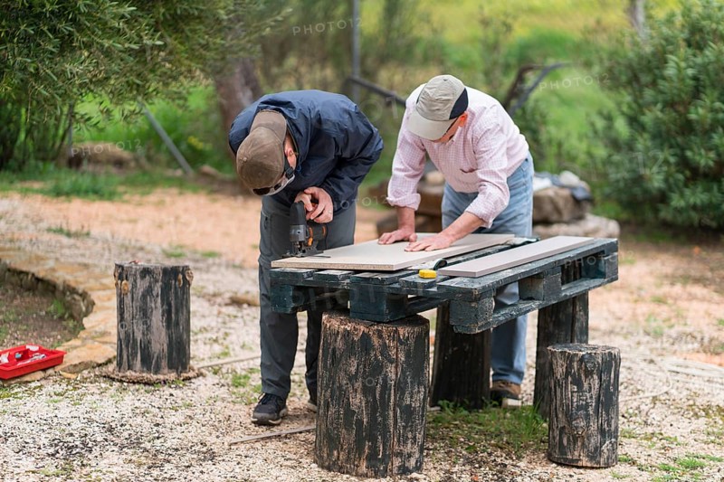 A senior father and his son bond while working on a carpentry project outdoors. Both are focused on cutting a wooden plank using a circular saw at a makeshift workstation under open skies