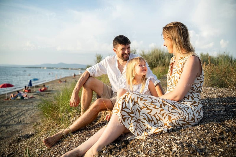 A young family sits on a sandy beach in Italy, enjoying a golden sunset. The parents lovingly interact with their daughter, creating cherished memories by the Mediterranean shore