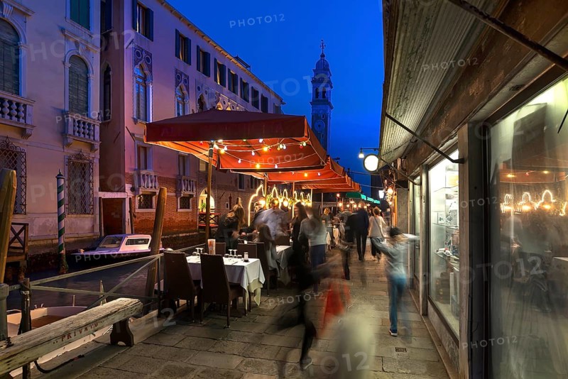 In the evening, lively restaurant on a canal with the tower of Santa Maria della Pietà in the back, Venice, Veneto, Italy