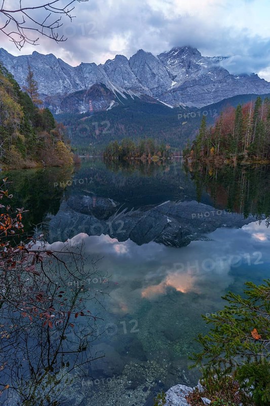 The serene waters of Eibsee capture the majestic Zugspitze peak, reflecting autumn foliage and rugged mountain peaks under a cloudy sky, creating a tranquil landscape