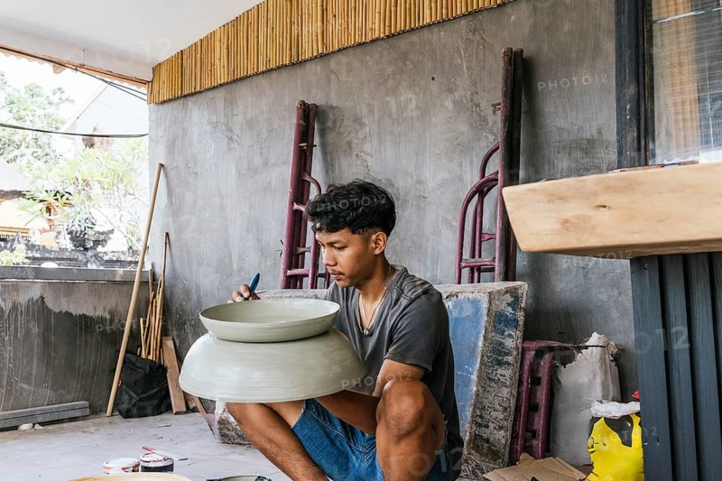 A Balinese artist intently paints a large sculpture in a bright studio. The space is adorned with bamboo and natural elements, enhancing the artistic ambiance