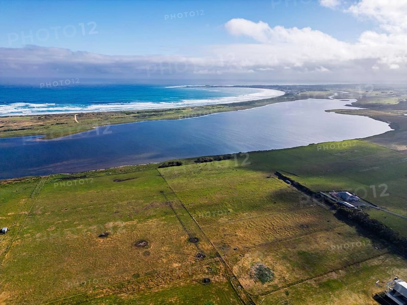 A stunning aerial view captures a coastal lake seamlessly blending into the ocean under a clear sky, with lush green fields surrounding the area, offering a peaceful landscape of Victoria, Australia