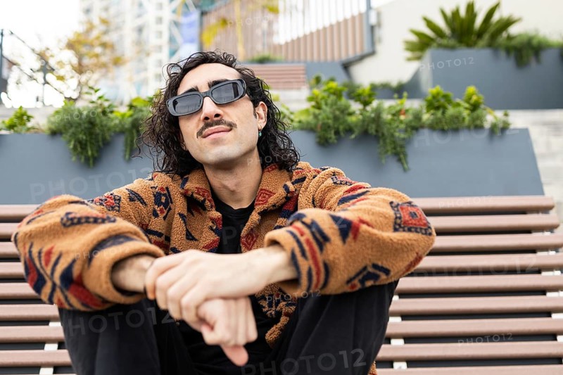Stylish man with sunglasses and patterned jacket sits relaxed on a bench in an urban setting The background features greenery and blurred city architecture, conveying a modern lifestyle vibe