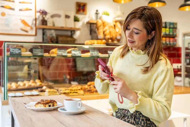Woman standing at a wooden counter, holding a cellphone, and capturing a flat white coffee and cinnamon roll for social media content in a modern bakery or cafe