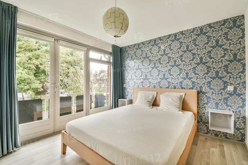 A cozy bedroom features a large bed with white bedding against a stunning blue floral wallpaper. Natural light floods through the large windows, creating a serene atmosphere