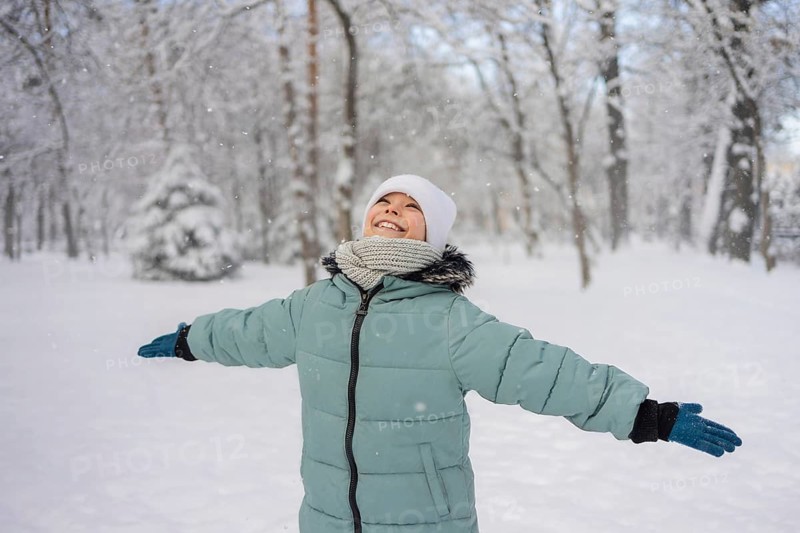A child in a green coat, white hat, and scarf joyfully stretches their arms in a snow-covered park. The serene snowy forest creates a winter wonderland backdrop