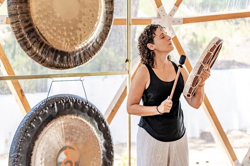 A female sound therapist performs a holistic session using a shamanic drum whit gongs standing behind her. The setup is in a serene outdoor retreat, promoting relaxation and wellness through sound therapy