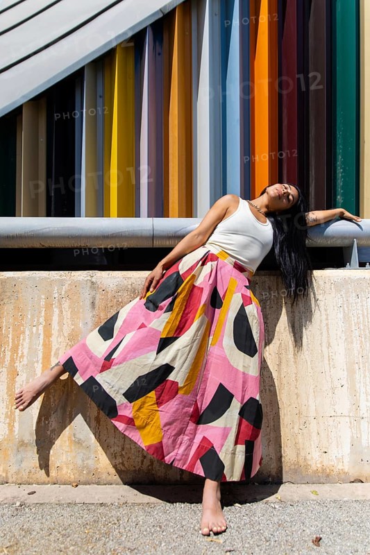 A woman in a vibrant, colorful outfit leans gracefully against a textured wall, showcasing a striking contrast with the modern architecture's clean lines and bold hues