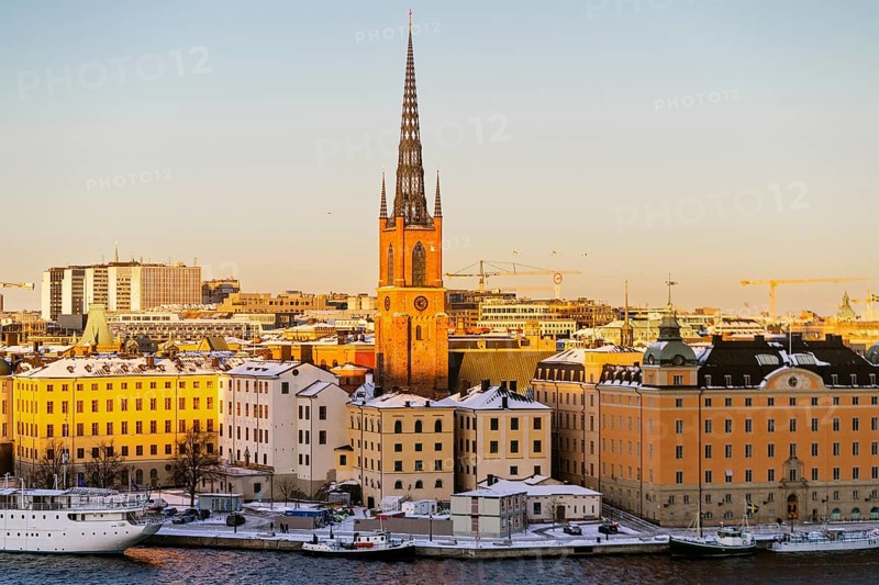 A winter view of Stockholm's historic waterfront, showcasing snow-dusted buildings and a prominent church spire under a golden sunset, highlighting the city's architectural beauty