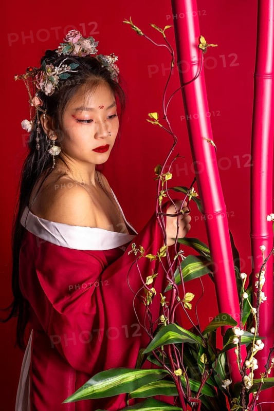 A woman in a red dress with a flower headpiece stands in front of a red background