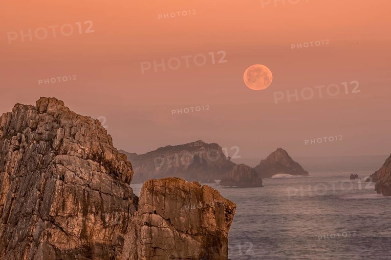 The full moon hovers above jagged rock formations along the Atlantic coast of Arnia Beach, Costa Quebrada, as the sky glows with soft sunrise hues in Cantabria
