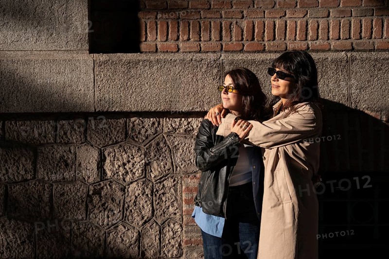 A multiethnic lesbian couple embraces warmly, basking in the sunlight against a brick and stone urban backdrop, symbolizing love and pride in diversity