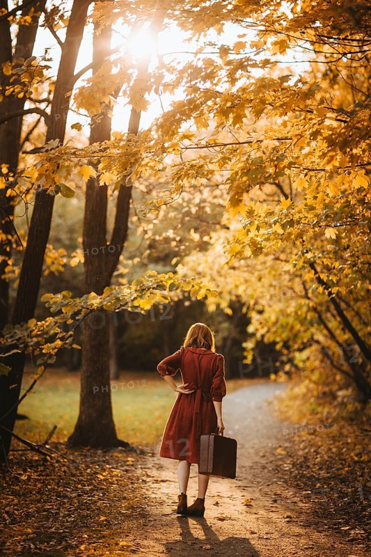 A woman in a red coat walks along a sunlit path in a forest during autumn in Quebec, Canada. The golden foliage and soft light create a serene and picturesque atmosphere