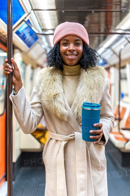 Smiling young black woman in winter coat holding a reusable coffee cup while riding a subway train