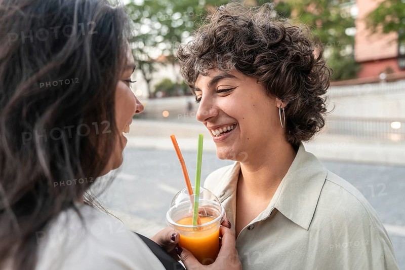 A joyful lesbian couple shares a refreshing drink outdoors, smiling warmly. They enjoy a leisurely moment in a charming urban setting, capturing a sense of love and friendship