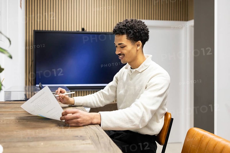A young professional sits at a wooden desk, reviewing documents with a pen in hand. The modern office setting, featuring a screen and stylish decor, reflects productivity and focus
