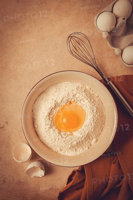 Food background, concept, plate with wheat flour, broken egg, whisk, kitchen towel, on a beige table, top view, rustic